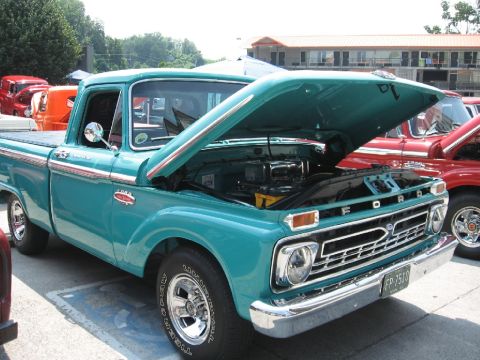 1966 Ford F-100 - A teal 1966 Ford F-100 pickup truck is displayed with its hood open, revealing the engine. Another red truck is visible to the right.