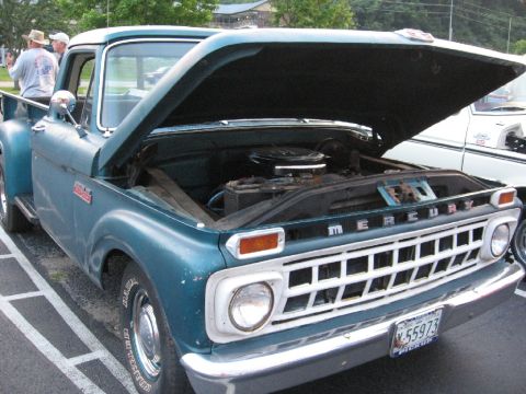 1966 Ford F-100 - A teal Mercury pickup truck is parked with its hood open, revealing the engine and battery. The grille spells out 'MERCURY'.