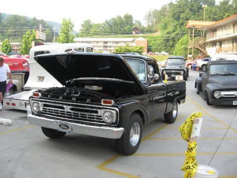 1966 Ford F-100 - A black 1966 Ford F-100 pickup truck is displayed with its hood open, alongside other vehicles.