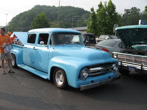 1966 Ford F-100 - A light blue pickup truck with four doors and a long bed is parked on asphalt, with trees and hills in the background.