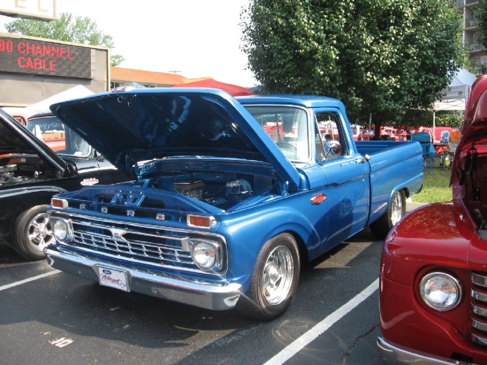 Blue pickup truck with hood open, parked next to a red truck. A sign in the background says '80 CHANNEL CABLE'