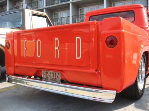 1966 Ford F-100 - Rear view of a bright orange Ford F-100 pickup truck, the iconic tailgate lettering and a chrome bumper.