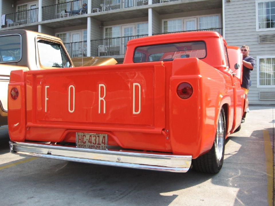 Rear view of an orange 1956 Ford F-100 pickup truck with chrome bumper and 'FORD' tailgate lettering. Another truck is partially visible to the left