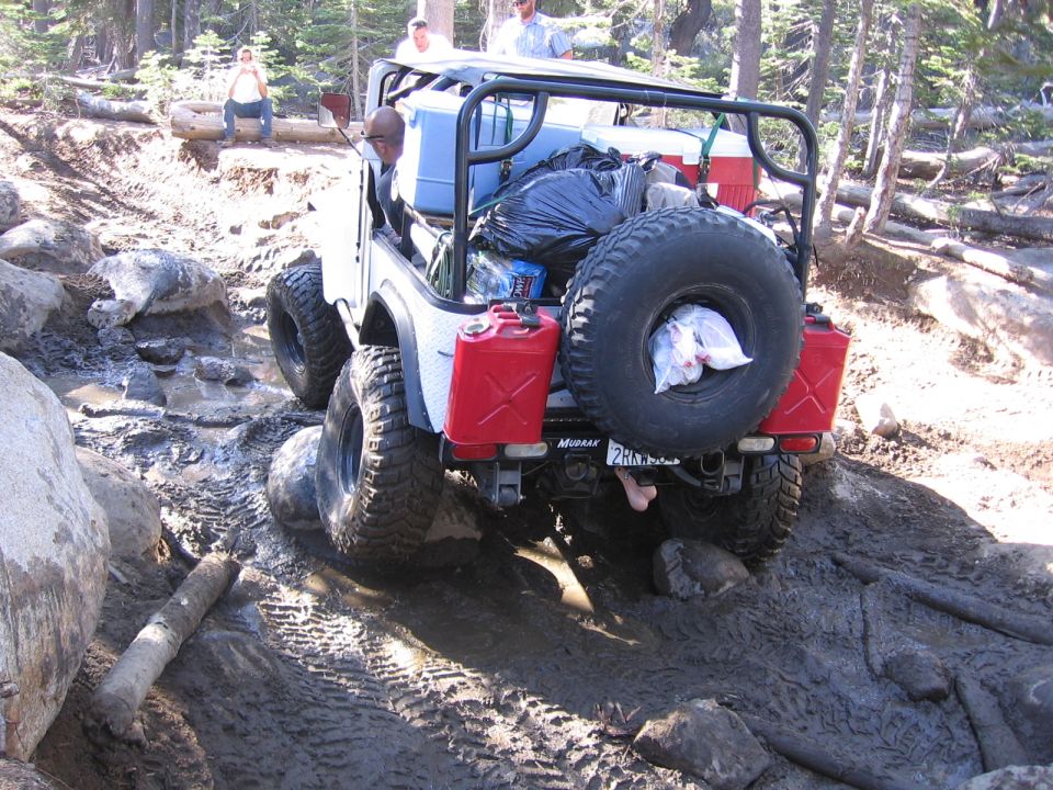 White off-road vehicle with a spare tire on the back, two red jerry cans, and camping gear, navigating a muddy trail