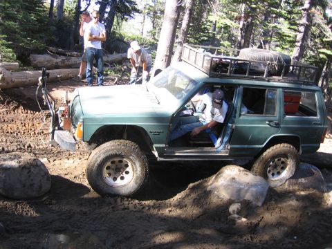 1989 Ford Bronco - A green Jeep Cherokee XJ is shown off-roading on a muddy, rocky trail with several people observing.