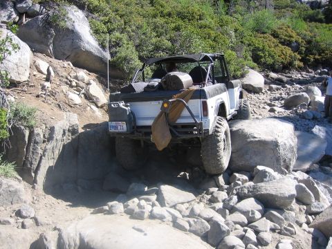 1989 Ford Bronco - Off-road SUV with soft top navigates a challenging rocky trail, showing its rear and side profile.