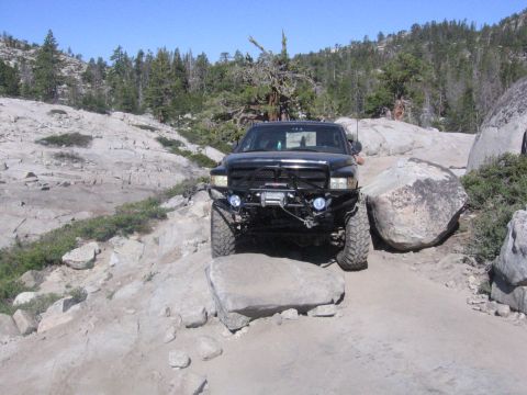 1989 Ford Bronco - Off-road pickup truck maneuvering over large rocks on a dirt path with trees and mountains in the background.