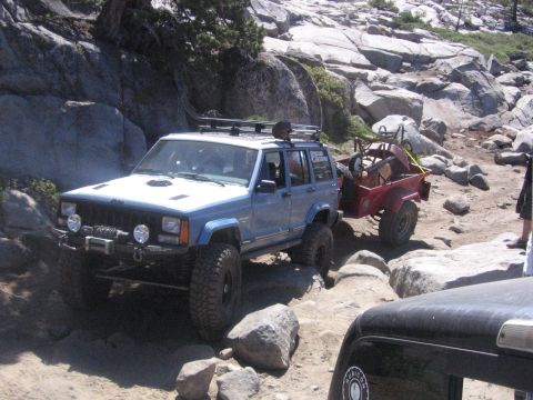 1989 Ford Bronco - A blue Jeep Cherokee XJ navigates a rocky off-road path while towing a red trailer.