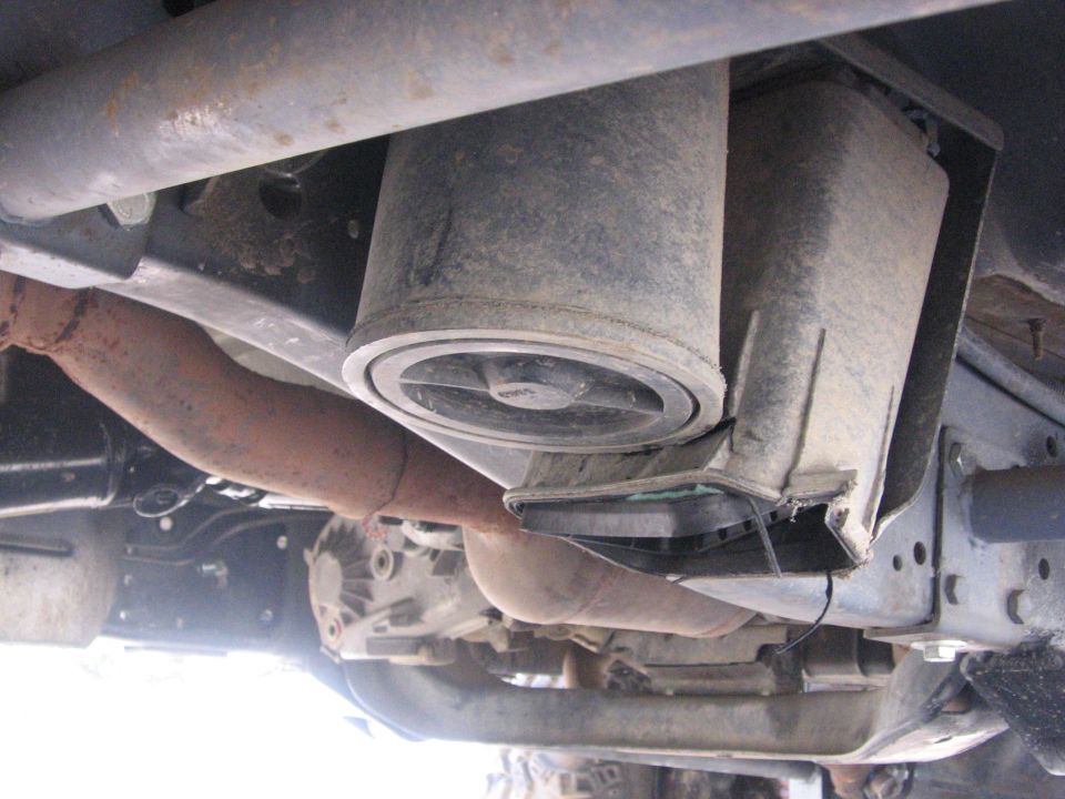 Underside view of a vehicle's suspension and exhaust system, showing a cylindrical component and rusty pipes