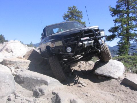 1989 Ford Bronco - Off-road truck with custom bumper and winch climbing over large rocks on a sunny day.