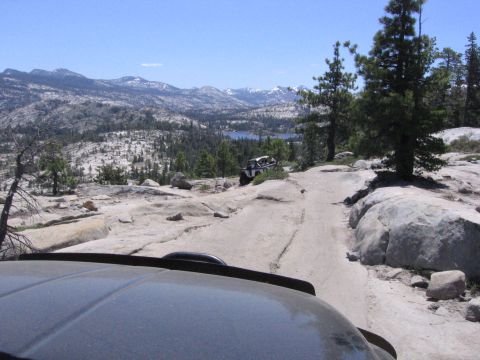 1989 Ford Bronco - Two off-road vehicles navigate a mountain trail with a scenic lake and snow-capped mountains in the background.