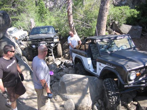 1989 Ford Bronco - Several off-road vehicles navigate a challenging rocky path in a wooded area.