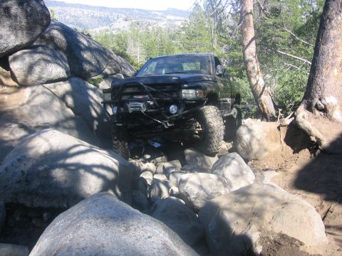 1989 Ford Bronco - A modified black pickup truck is shown traversing a challenging rocky terrain in a wooded area.