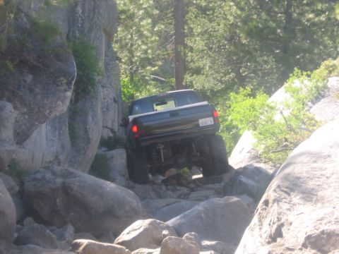 1989 Ford Bronco - A dark pickup truck with off-road modifications is seen from the rear, driving over large rocks on a trail.