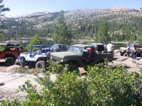 1989 Ford Bronco - Multiple off-road vehicles are parked on a dirt lot with green foliage and hills in the background.