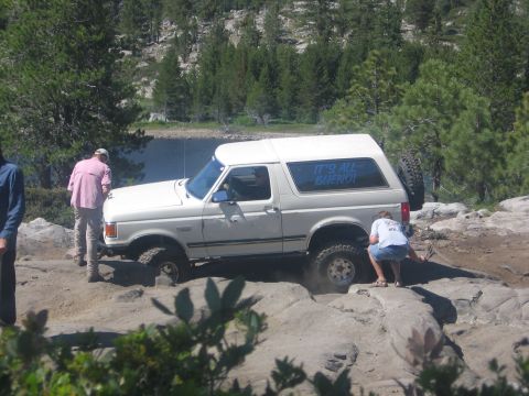 1989 Ford Bronco - A white 1989 Ford Bronco is parked on a rocky surface, with trees and another vehicle in the background.