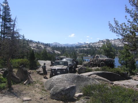 1989 Ford Bronco - A group of off-road vehicles are parked on a rocky terrain near a lake on a sunny day.