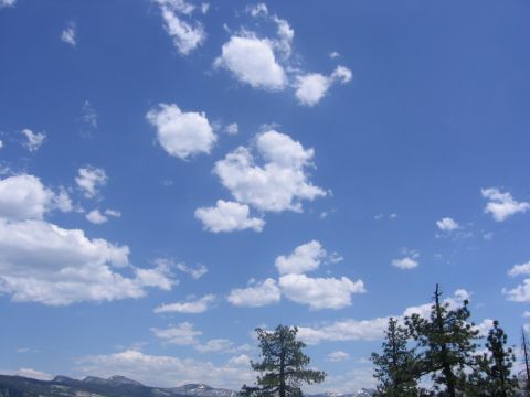 1989 Ford Bronco - A scenic view of a bright blue sky dotted with fluffy white clouds, with mountains and trees in the distance.