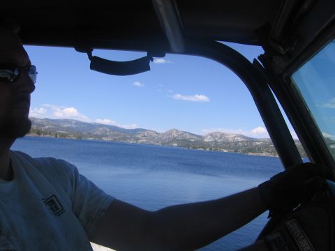 1989 Ford Bronco - Man drives a vehicle with a roll cage, looking out at a scenic lake and mountain landscape on a sunny day.