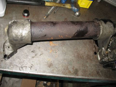 1995 Ford C-350 (Centurion Conversions) - A removed steering column laid on a scarred metal workbench, its rusty tube running between the cast upper housing with gear and a cast alu…