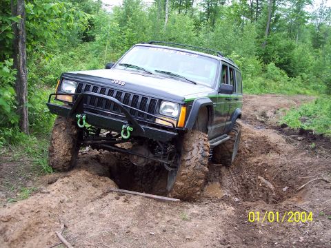 1979 Ford Bronco - A modified Jeep Cherokee XJ is shown tackling a challenging muddy terrain with large off-road tires.