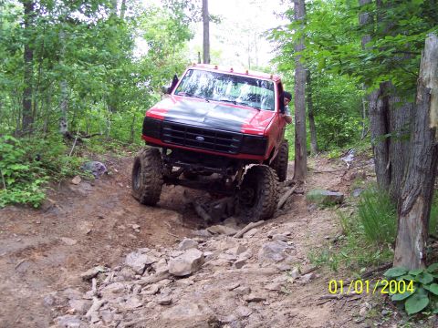 1979 Ford Bronco - A red lifted truck tackles a challenging off-road course through a wooded area.