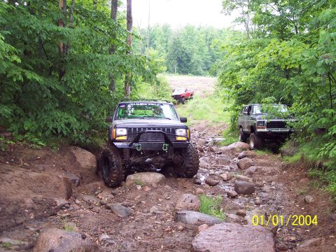 1979 Ford Bronco - Two off-road vehicles navigate a challenging, rocky path through a dense forest.
