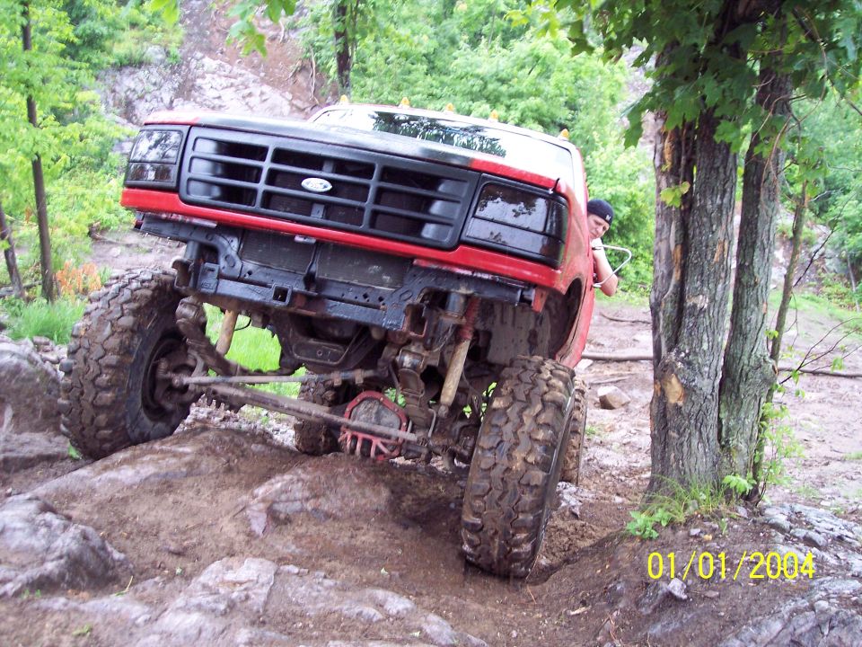 Red truck with large tires climbing over rocks and dirt. Person visible behind the wheel well