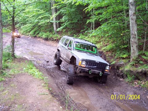 1979 Ford Bronco - A Jeep Cherokee XJ navigates a challenging muddy trail in a wooded area, with another vehicle visible in the distance.
