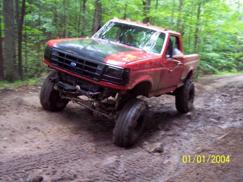 1979 Ford Bronco - This heavily modified red pickup truck navigates a muddy off-road path through a wooded area.