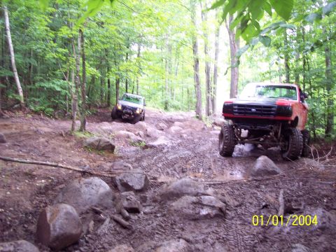 1979 Ford Bronco - Two off-road vehicles navigate a challenging, wooded path with large rocks and mud.