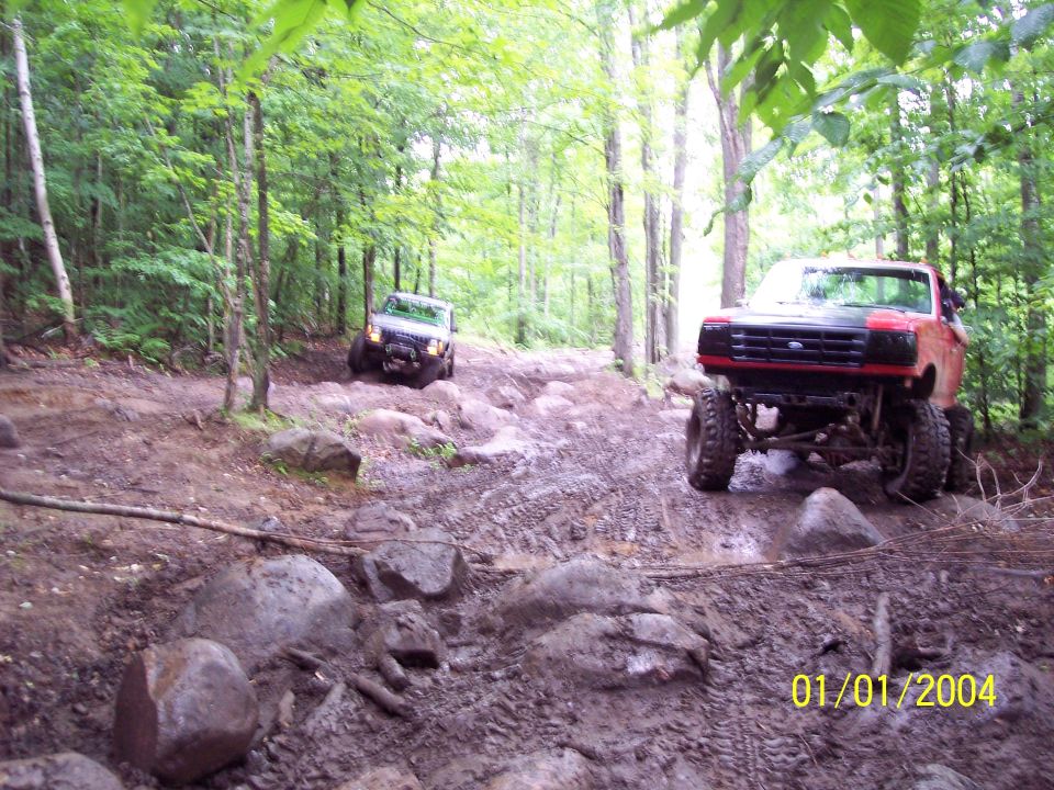 Red Ford truck and a silver SUV driving on a rocky, muddy trail in the woods