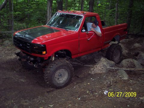 1979 Ford Bronco - A red pickup truck with a black hood and lifted suspension navigates a rocky, wooded trail.