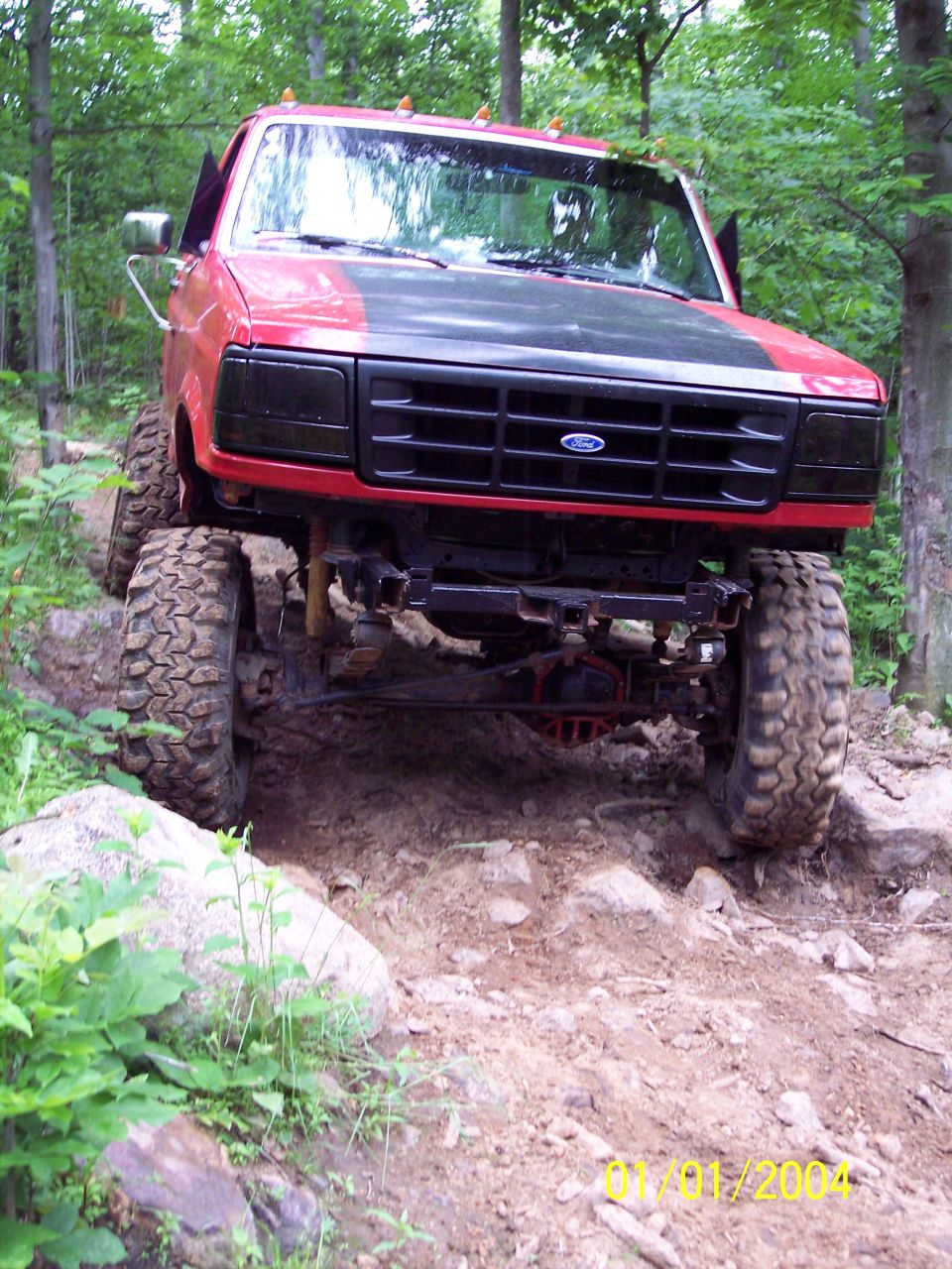 Red Ford truck with black grille and hood, lifted suspension, off-roading on a rocky trail