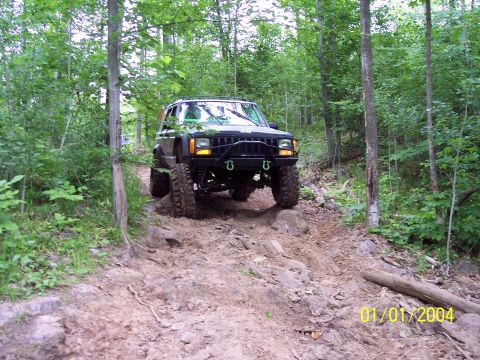 1979 Ford Bronco - A modified black Jeep Cherokee XJ navigates a challenging, rocky off-road trail surrounded by dense forest.