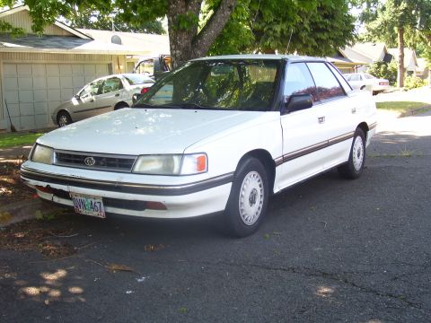 1991 Ford Bronco - This white sedan, likely from the early 1990s, is parked on a residential street next to a garage.