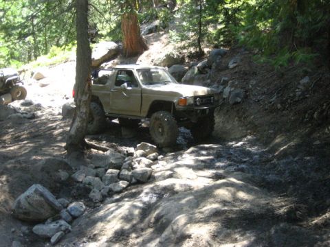 1989 Ford Bronco - Modified off-road vehicle tackling a challenging rocky path in a wooded area.