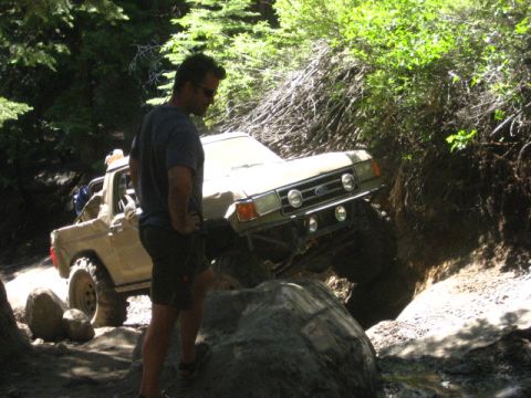 1989 Ford Bronco - A man watches a tan Ford Bronco navigate a challenging rocky incline on a wooded trail.