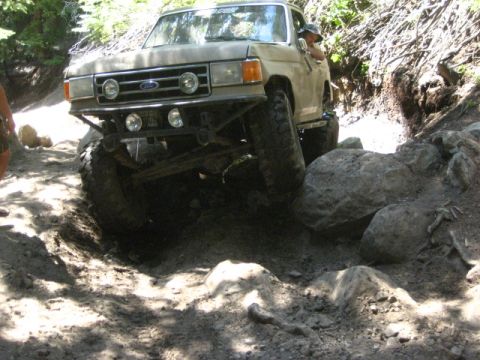 1989 Ford Bronco - Tan Ford Bronco tackles a challenging rocky off-road path with its front-left tire elevated.