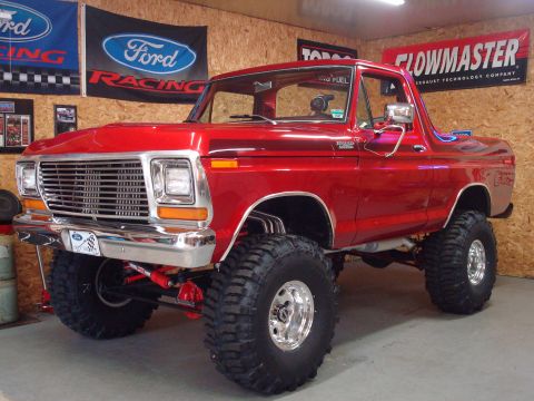 1978 Ford Bronco - Front three-quarter view of a glossy red late-1970s Ford Bronco on a tall suspension lift, sitting in an OSB-walled shop hung with Ford Rac…