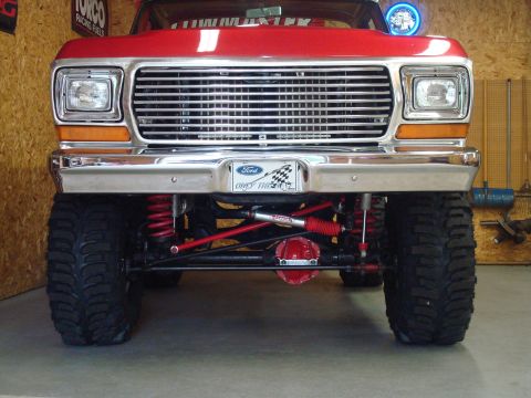 1978 Ford Bronco - Head-on view of a lifted red Ford 4x4 in a shop, showing the chrome egg-crate grille, rectangular sealed-beam headlights, amber turn signal…