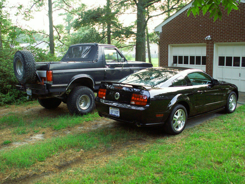 Black lifted Ford Bronco and black Mustang coupe parked on grass by brick garage