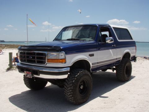 1996 Ford Bronco - A lifted 1996 Ford Bronco sits on the sand near the water under a bright blue sky.