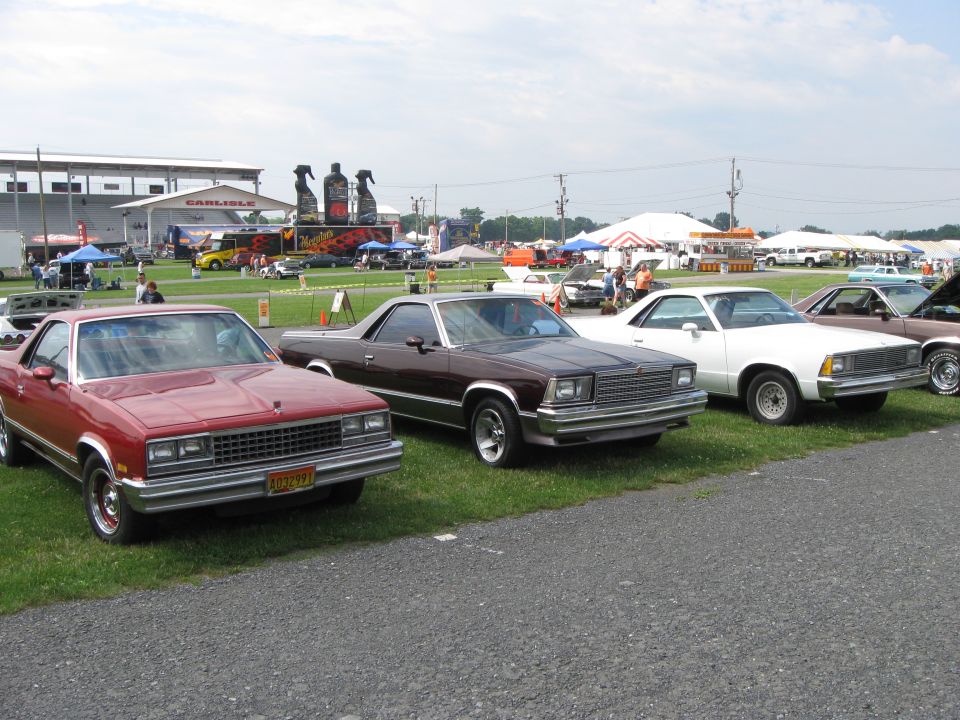Red El Camino in foreground, brown El Camino next, white El Camino beyond, parked on grass with tents and buildings in background