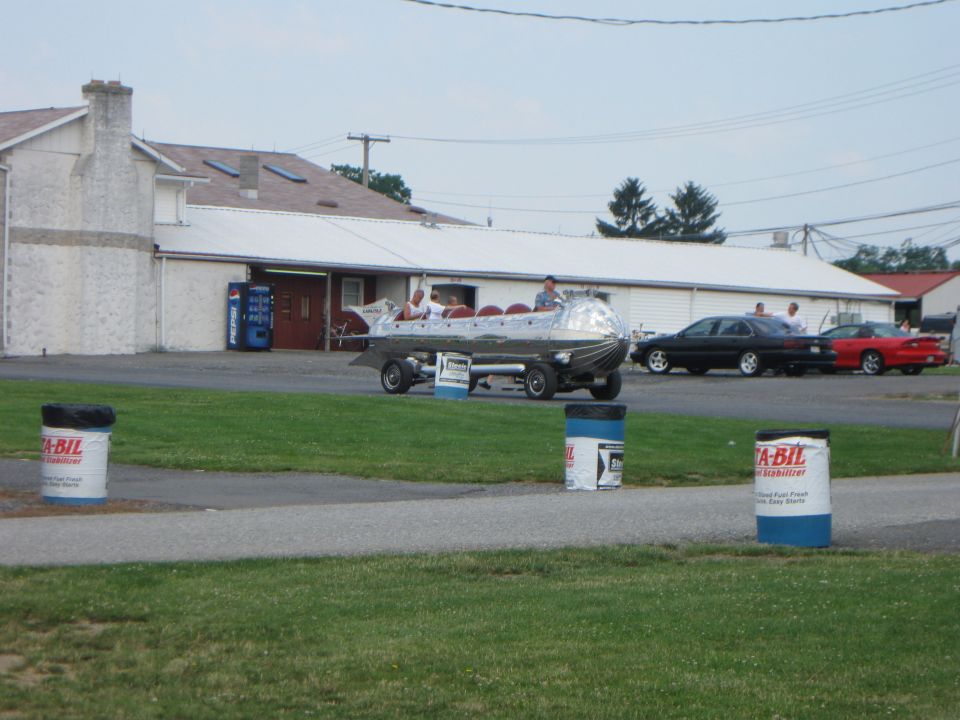 Silver novelty vehicle shaped like a rocket on a trailer, with people inside. Two cars are parked behind it