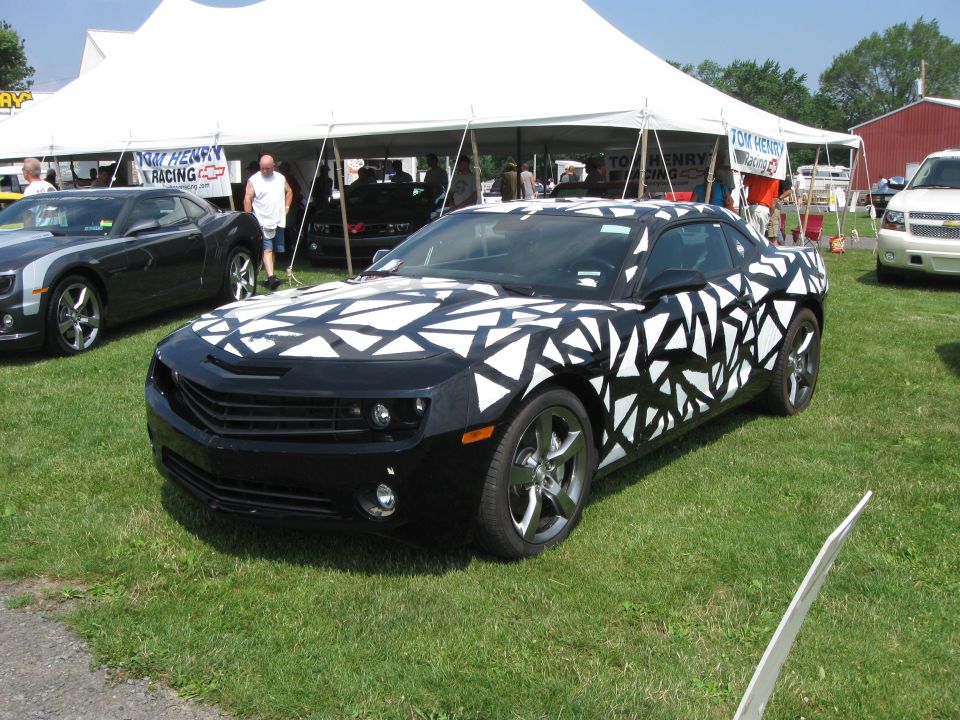 Black Chevrolet Camaro with a white geometric pattern wrap parked on grass under a tent