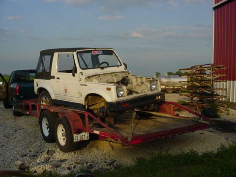 1992 Ford Bronco - A white Suzuki Samurai is loaded onto a trailer, with its hood open, parked next to a dark pickup truck.