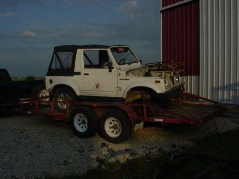 1992 Ford Bronco - Suzuki Samurai on a trailer in front of a building with red and silver siding.