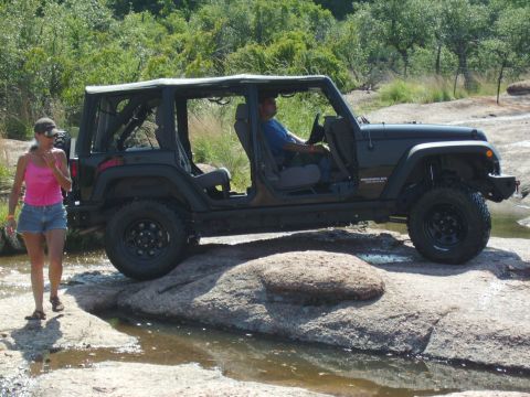 2008 Jeep Wrangler - A black Jeep Wrangler is driven over rocks in a stream, with a woman standing nearby.