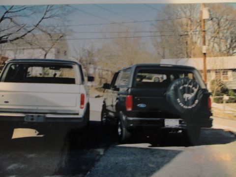 1995 Ford Bronco - Two Ford Broncos, one black and one white, are parked on a residential street. The black Bronco is in the foreground, showing its rear and…